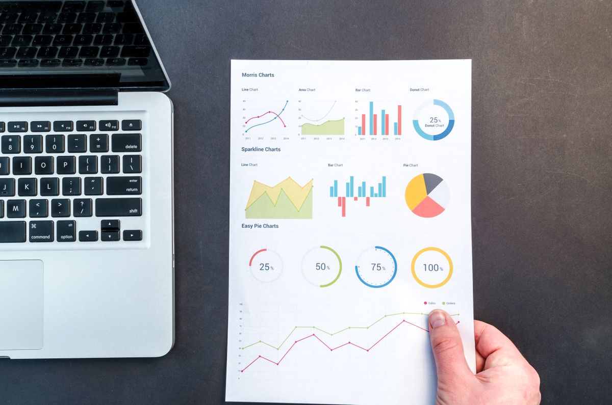 A hand holding a printed page of charts and graphs next to a laptop